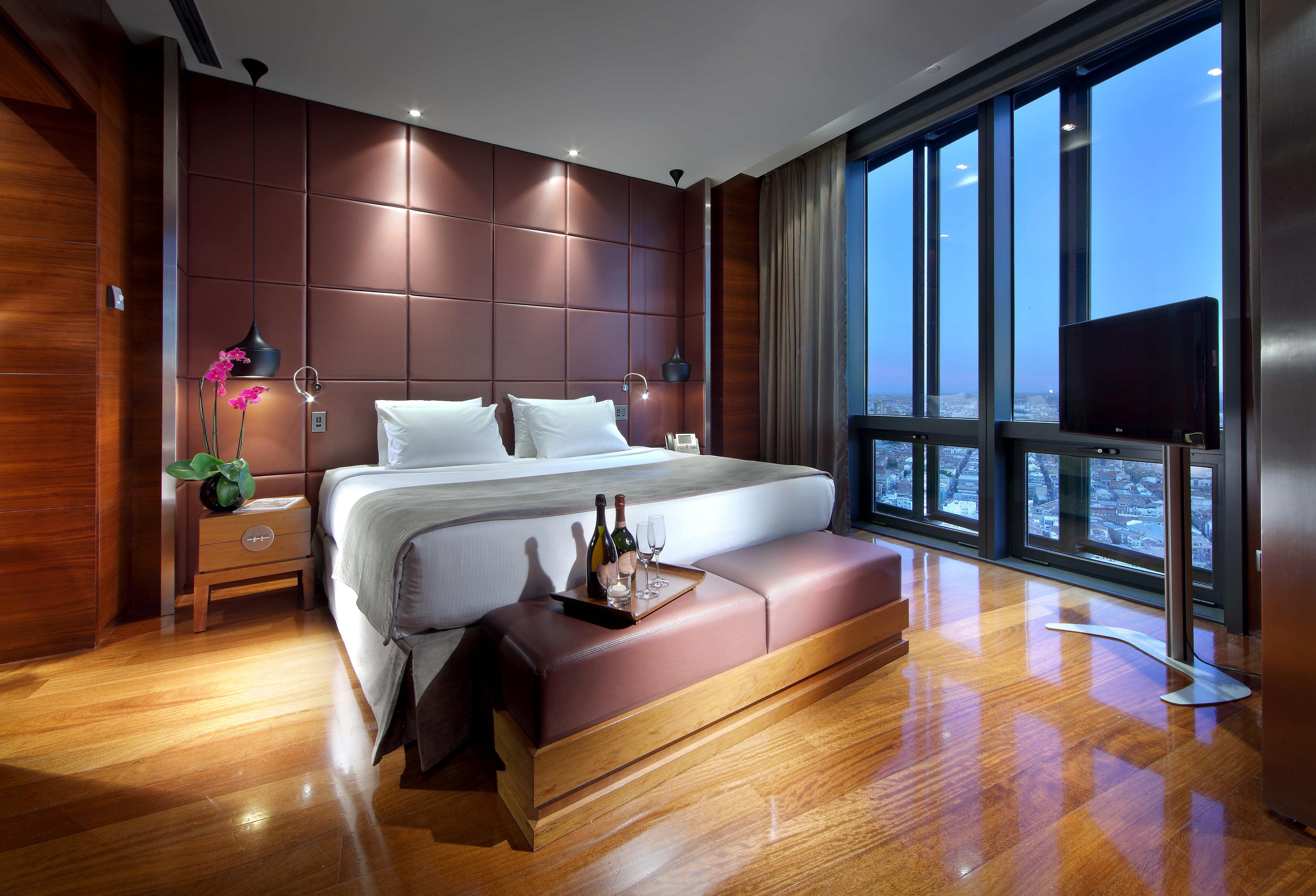 Eurostars Madrid Tower Hotell Eksteriør bilde The photo shows a modern hotel room with a luxurious design. It features a large bed with white linens and plush pillows, positioned against a stylish brown accent wall. There are bedside tables on each side of the bed, each equipped with a lamp. In 