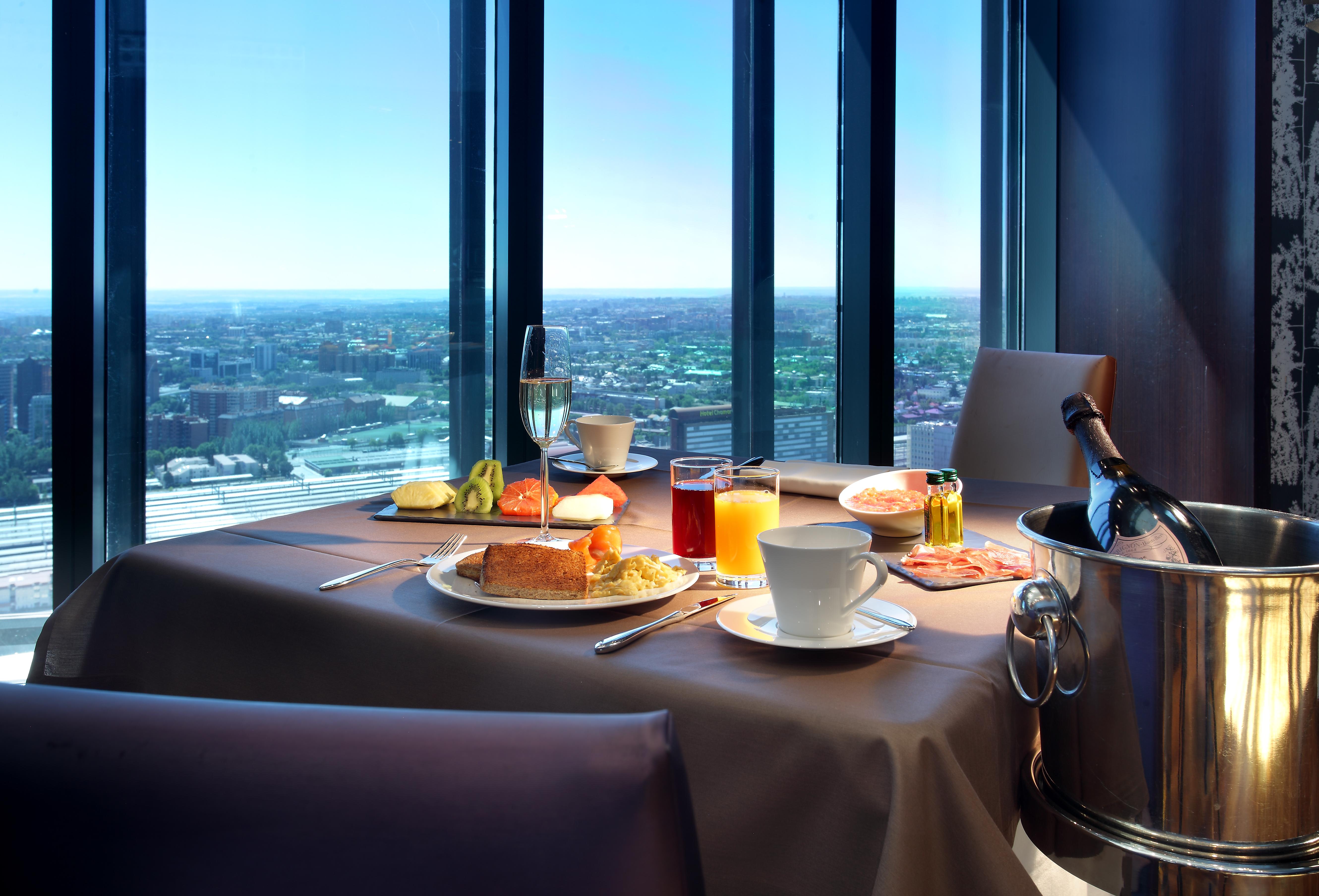 Eurostars Madrid Tower Hotell Eksteriør bilde The photo shows a beautifully set table with a scenic view through large windows. On the table, there are various food items, including a breakfast plate with items like eggs, pastries, and fruit. There are drinks, including a cup of coffee, a glass 