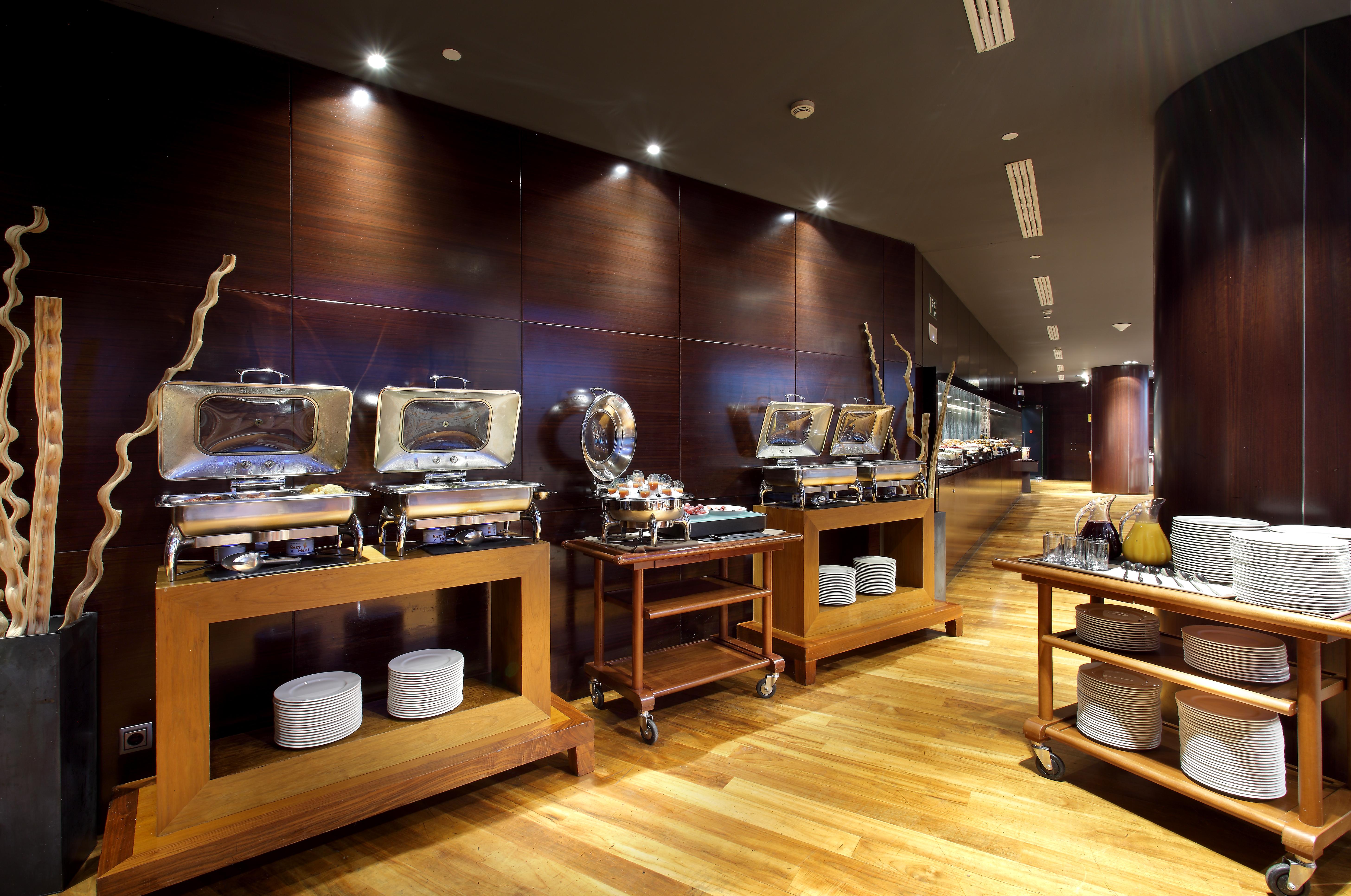 Eurostars Madrid Tower Hotell Eksteriør bilde The photo shows a modern restaurant buffet setting. There are several food warmers placed on wooden tables, showcasing a variety of dishes. The walls have a dark wood finish, and the lighting is soft and ambient, creating an elegant atmosphere. In fr