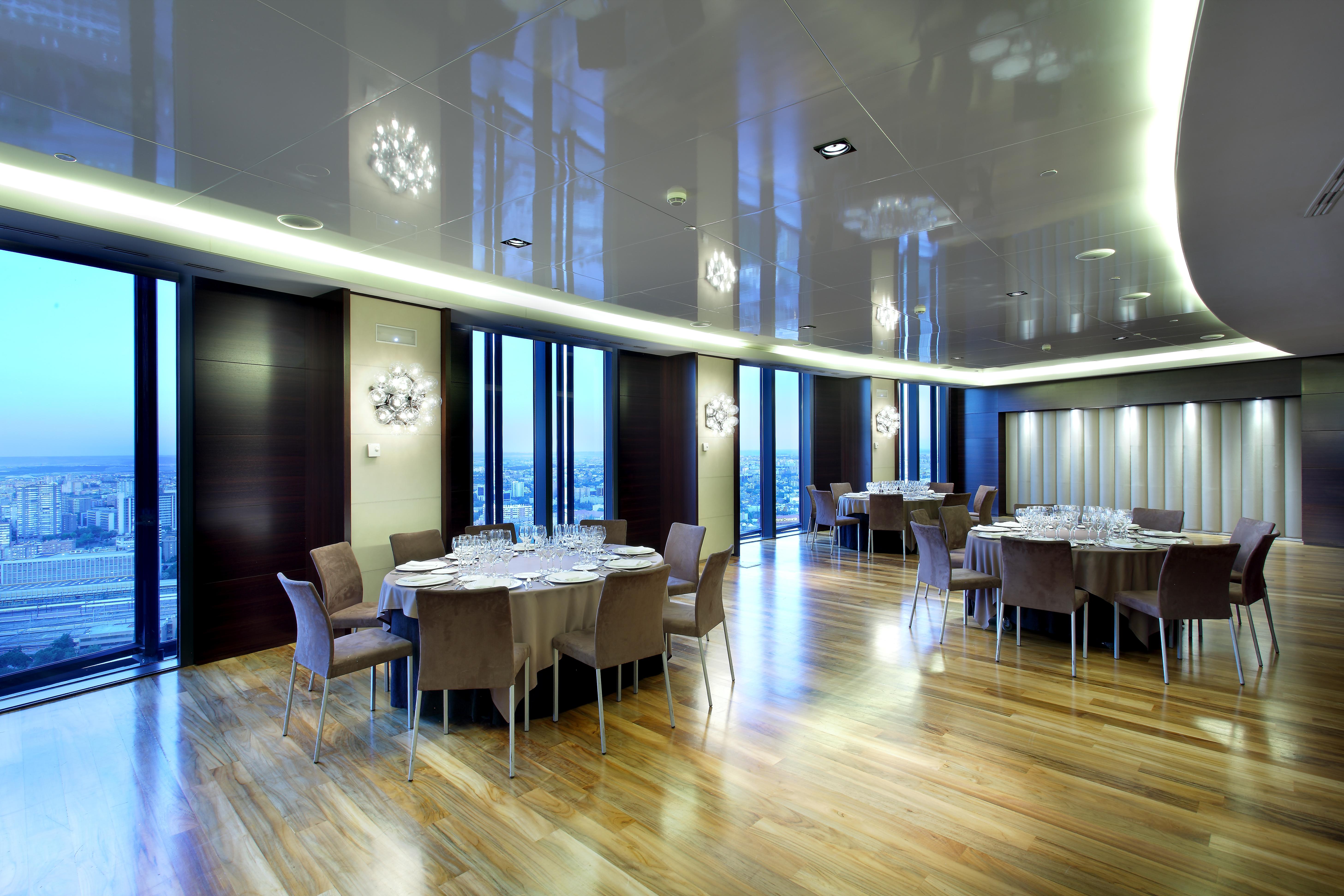 Eurostars Madrid Tower Hotell Eksteriør bilde The photo depicts a modern dining room or banquet hall with large windows that provide a view of the outside. The room features elegant wooden flooring and contemporary furniture, including round tables set with plates, glasses, and cutlery. Various 