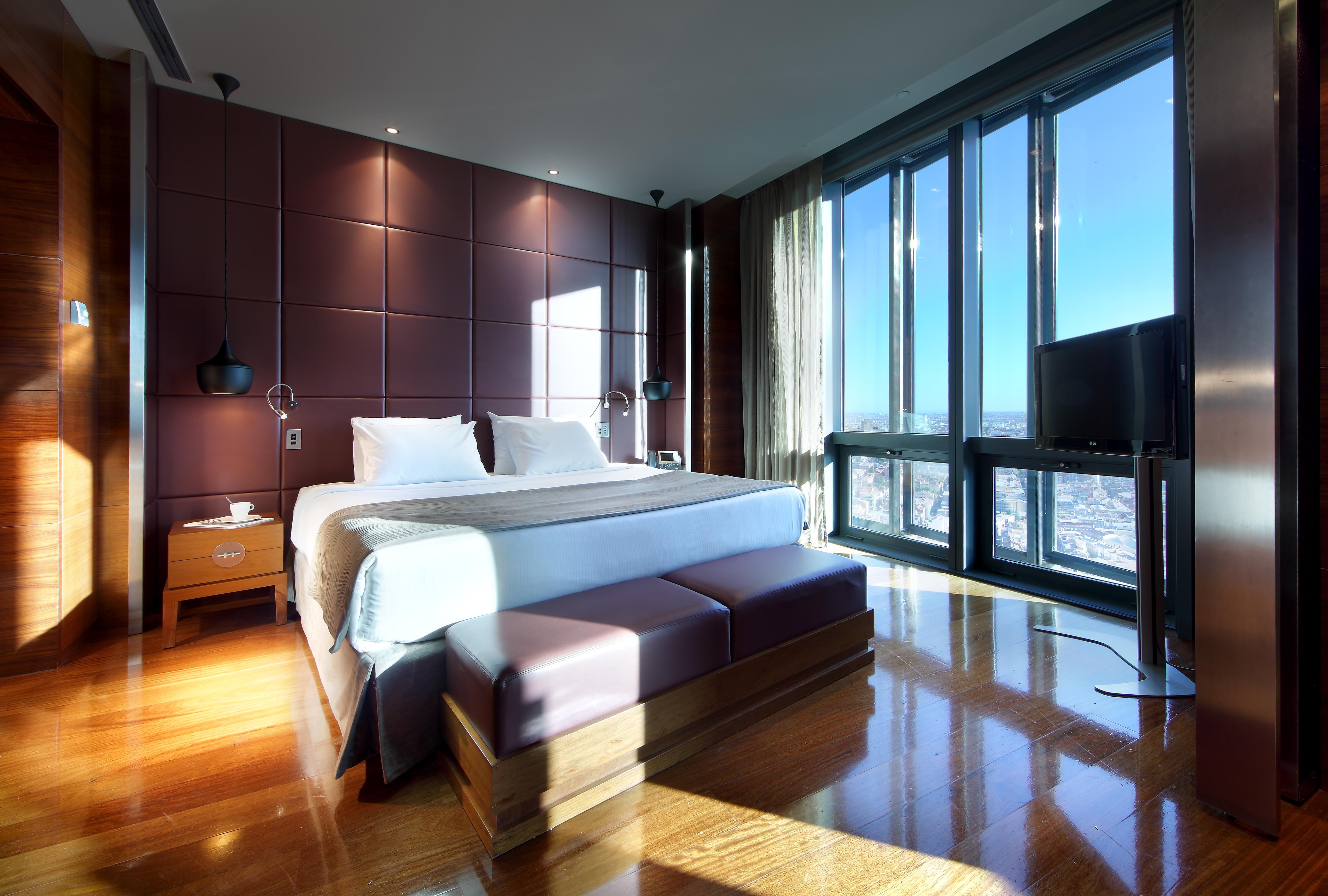 Eurostars Madrid Tower Hotell Eksteriør bilde The photo shows a modern hotel room with sleek design features. It includes a large bed with white bedding, positioned against a wall with dark, textured panels. On either side of the bed, there are bedside tables with lamps. A stylish bench sits at 