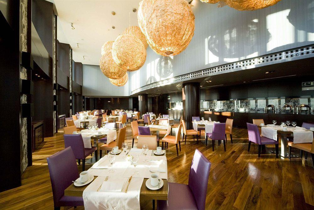 Eurostars Madrid Tower Hotell Restaurant bilde The photo shows a modern restaurant interior featuring elegant decor. The dining area includes tables set for guests with white tablecloths, plates, and cutlery. The seating consists of a mix of wooden chairs, some upholstered in purple fabric. The l