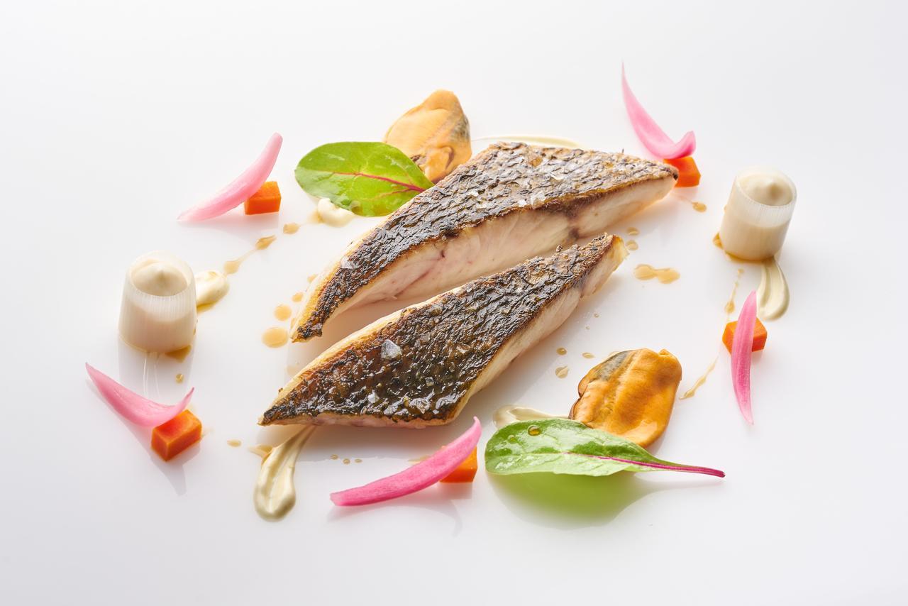 Eurostars Madrid Tower Hotell Eksteriør bilde The image depicts a beautifully plated dish featuring two pieces of fish fillet, likely a type of white fish, arranged artfully on a white plate. The fish has a crispy skin and is cooked to a tender texture. Surrounding the fish are various garnishes