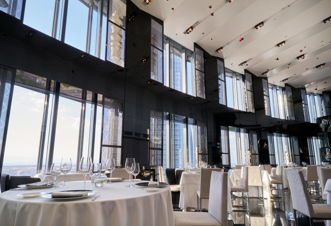 Eurostars Madrid Tower Hotell Eksteriør bilde The photo depicts an elegant restaurant interior characterized by sleek, modern design. It features large floor-to-ceiling windows that allow abundant natural light to fill the space and provide a panoramic view of the outside. The dining area includ