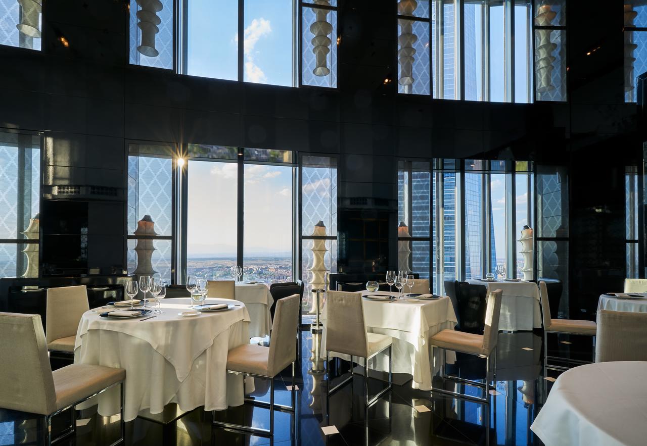 Eurostars Madrid Tower Hotell Eksteriør bilde The photo features an elegant restaurant interior with large windows that provide a panoramic view of the city skyline. The tables are elegantly set with white tablecloths and fine dining tableware, including glasses and plates. The decor appears mod