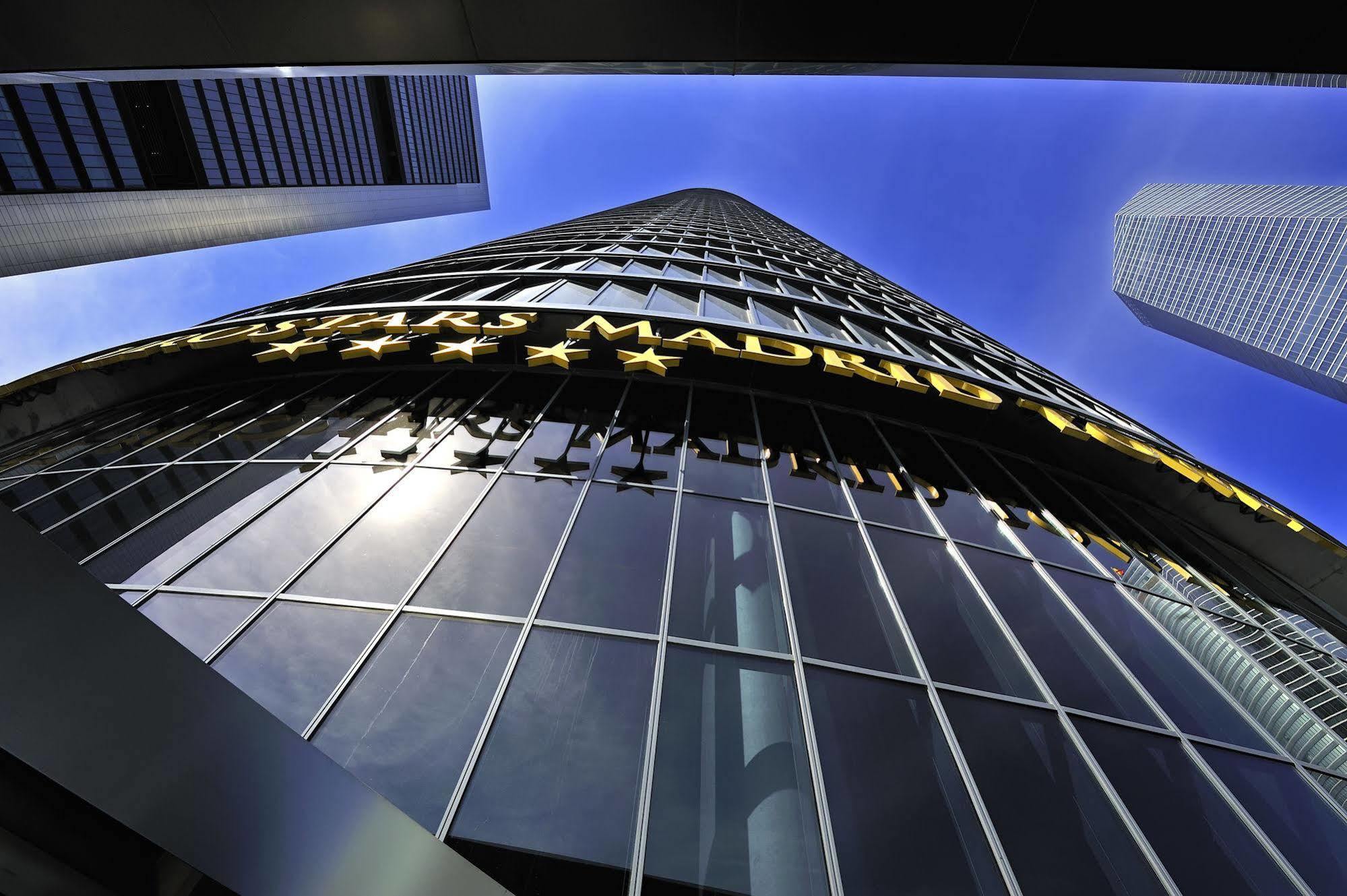 Eurostars Madrid Tower Hotell Eksteriør bilde The photo shows a tall modern building reaching towards the sky. The perspective is from the ground looking upward, highlighting the building's height and glass facade. The building features a large sign that reads "MADRID" in bright yellow letters, 